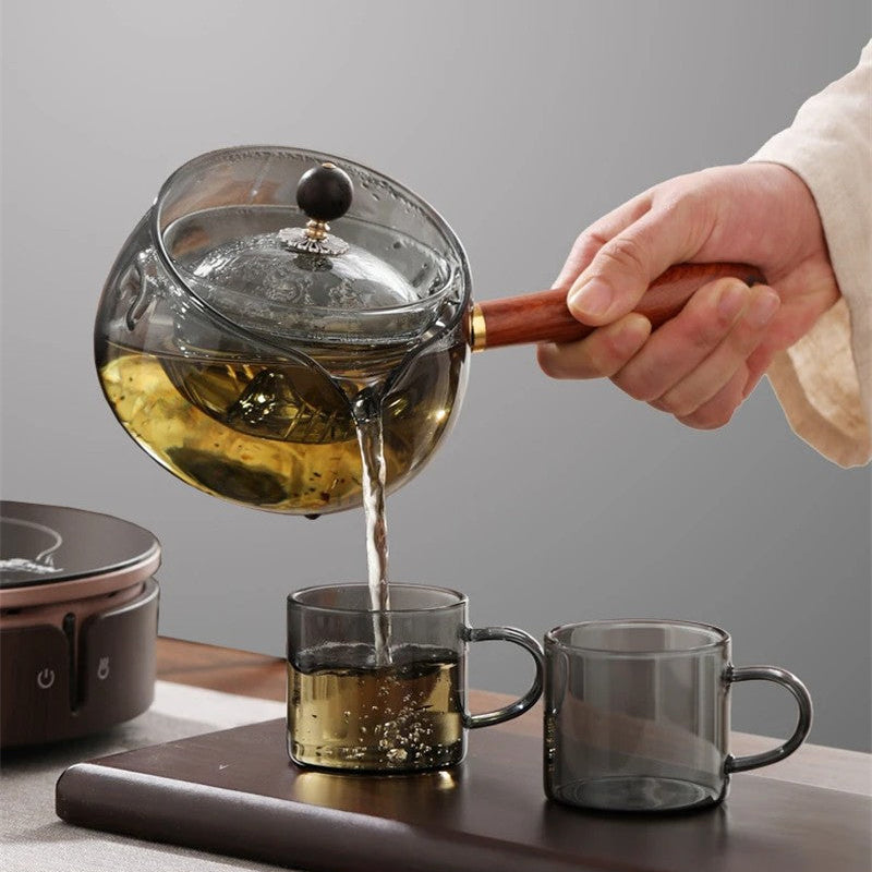 Person pouring tea from a glass teapot into two cups on a wooden tray with a gray background.